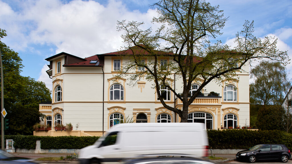 Außenansicht einer großen, cremefarbenen Altbauvilla mit Satteldach und Bäumen davor, mit einem vorbeifahrenden weißen Lieferwagen.