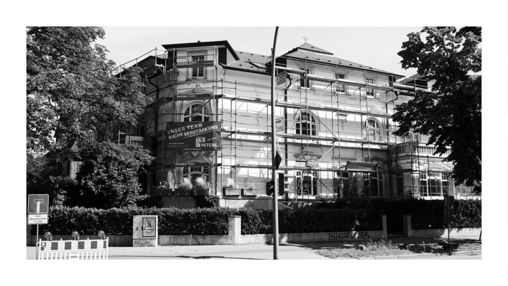 Schwarz-Weiß-Foto desselben Altbaus, umgeben von Gerüsten und Bauplanen, während der Sanierungsarbeiten.