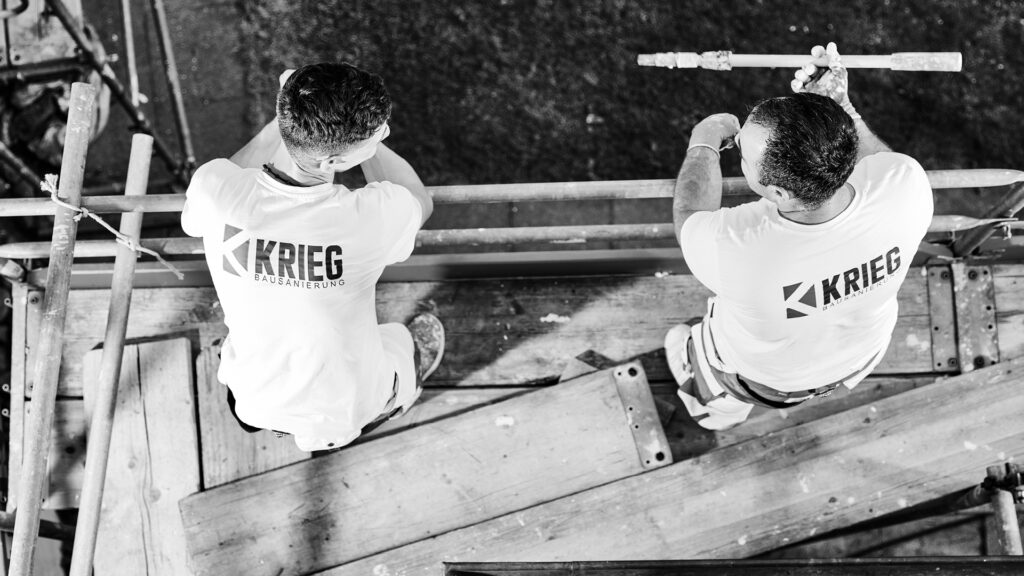 Schwarz-Weiß-Foto aus der Vogelperspektive, das zwei Handwerker der Krieg Bausanierung in T-Shirts mit Logo zeigt, die auf einem Baugerüst arbeiten.