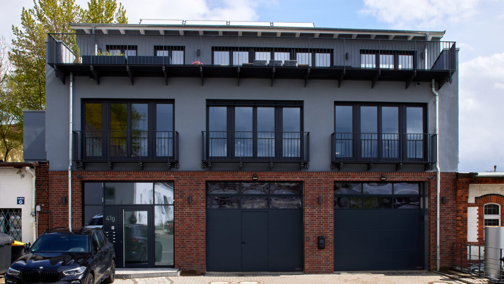 Moderne Gebäudefront mit dunkelgrauer Fassade, großen Fensterfronten und Garagentoren im Erdgeschoss der Produktionshalle sowie Balkonen mit Metallgeländern.