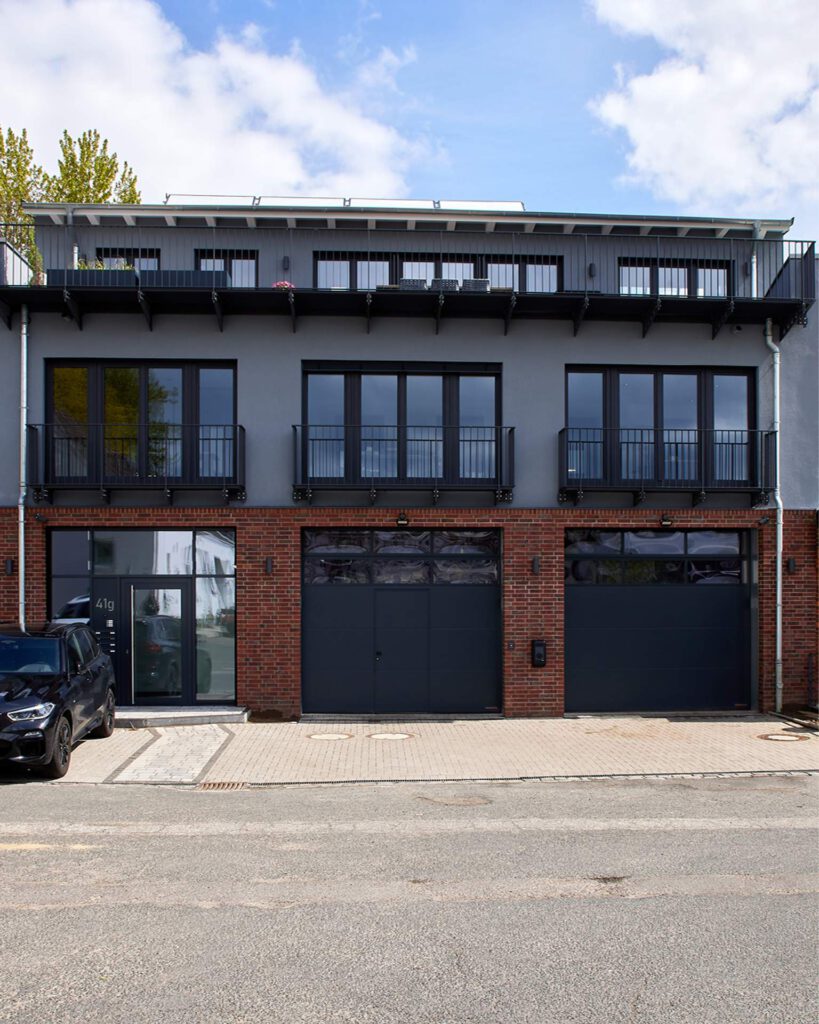 Moderne Gebäudefront mit dunkelgrauer Fassade, großen Fensterfronten und Garagentoren im Erdgeschoss der Produktionshalle sowie Balkonen mit Metallgeländern.