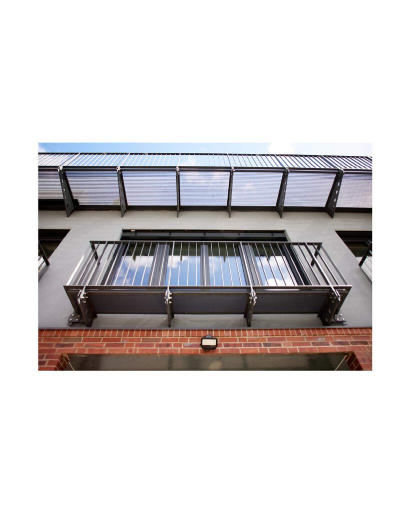 Nahaufnahme eines Balkons mit filigraneam schwarzem Metallgeländer vor einer hellgrauen WDVS-gedämmten Fassade unter blauem Himmel.