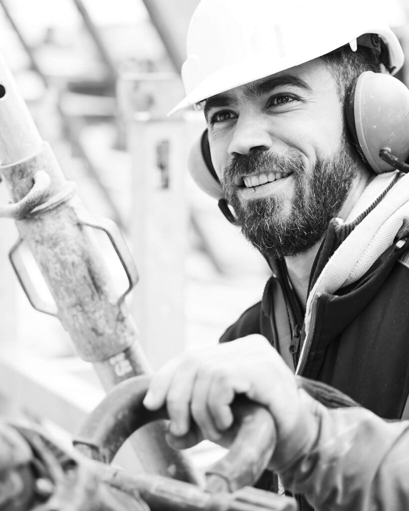 Lächelnder Bauhandwerker mit Schutzhelm und Gehörschutz bei der Arbeit an einer Stahlbetonkonstruktion – Sinnbild für Präzision und Teamgeist.