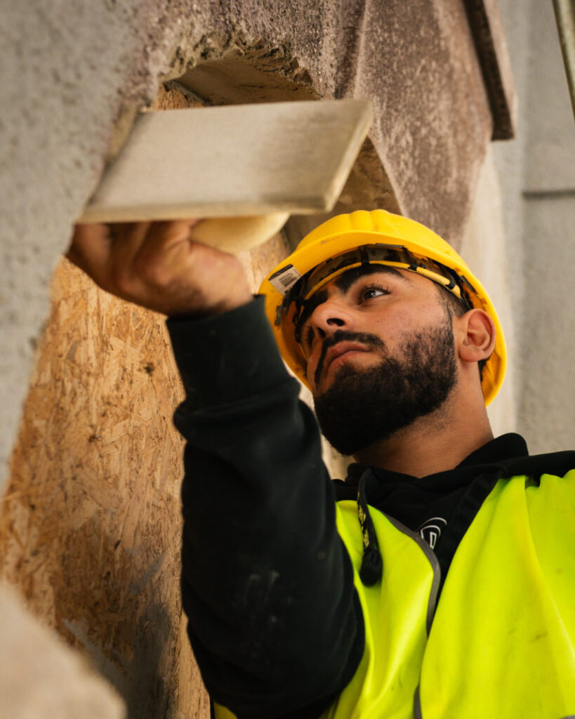 Mitarbeiter von Krieg bausanierung in gelber Warnweste und Helm schaut fokussiert auf die Spachtel in seiner Hand, mit der er eine Hauswand abbzieht.