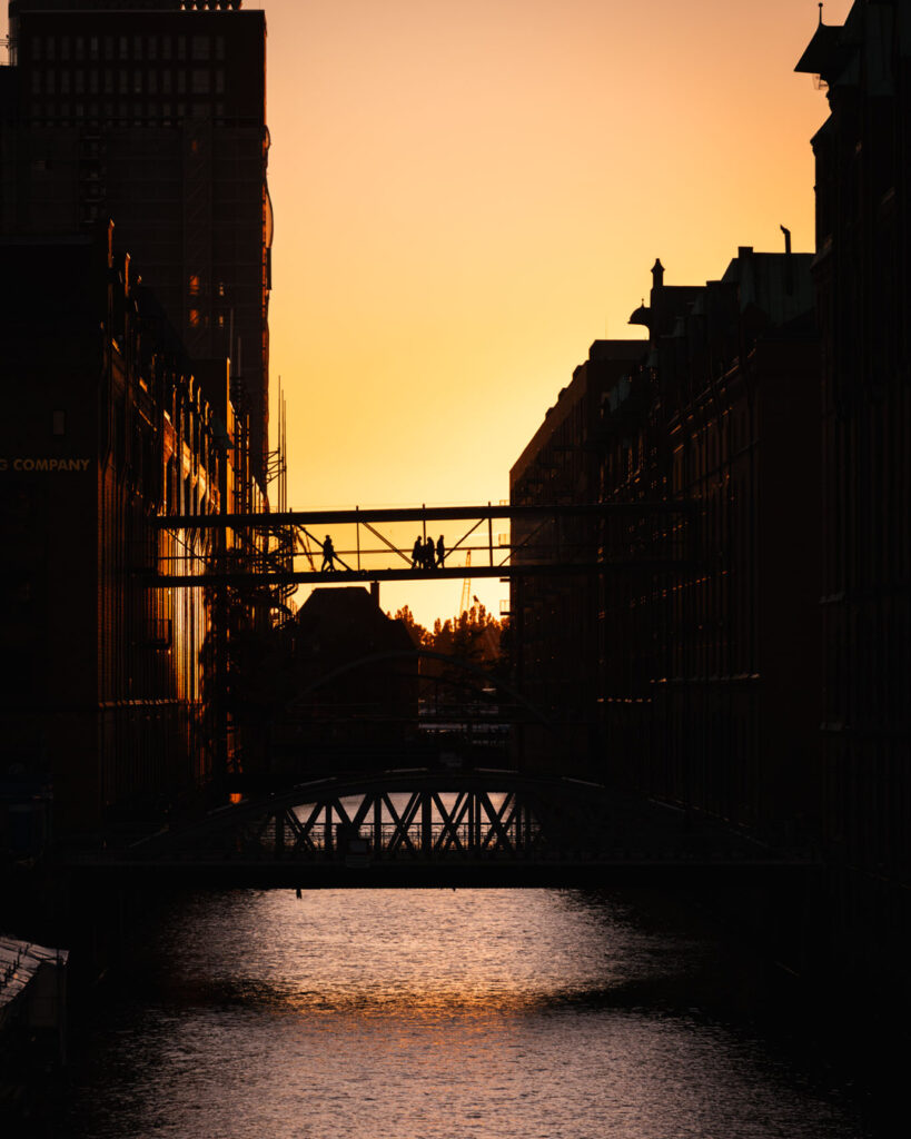 Panoramasicht Hamburg Speicherstadt bei Sonnenuntergang: Eine weite Perspektive des historischen Lagerhauskomplexes mit Backsteingebäuden, dem Fleet, dem Kehrwiedersteg und der charakteristischen Brücke mit Passanten im Gegenlicht.