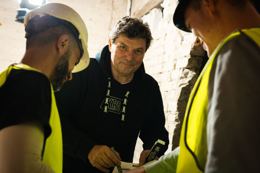 Experten der Krieg Bausanierung bei der Begutachtung einer Baustelle. Ein Bauleiter im schwarzen Hoodie bespricht technische Details mit Handwerkern in Sicherheitswesten vor einer freigelegten Ziegelwand. SEO-Fokus: Professionelle Bausanierung, Altbausanierung und Baustellenbesprechung durch Fachpersonal.