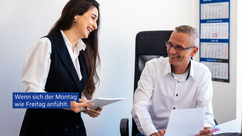 Zwei lachende Kollegen des Teams KRG im Büro besprechen Dokumente mit dem Text „Wenn sich der Montag wie Freitag anfühlt“.
