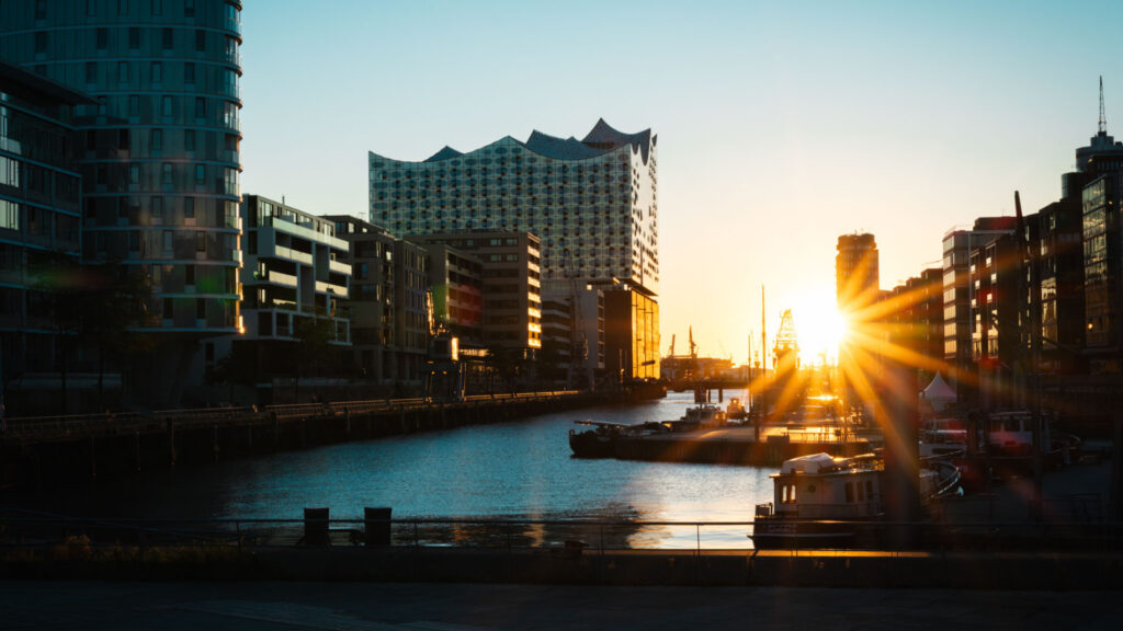 Sonnenuntergang in der Hamburger HafenCity mit Blick auf das moderne Konzerthaus Elbphilharmonie an einem Kanal, im Vordergrund Schiffe und Boote vor moderner Architektur im warmen Gegenlicht.