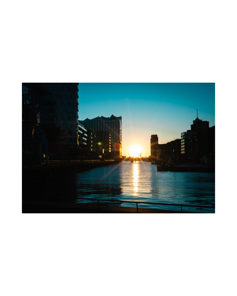 Hamburger Hafenbecken in der HafenCity bei Sonnenuntergang mit tiefstehender Sonne, starker Lichtreflexion auf dem Wasser, der Elbphilharmonie Silhouette und blauem Himmel im Hochformat mit weißem Rahmen.