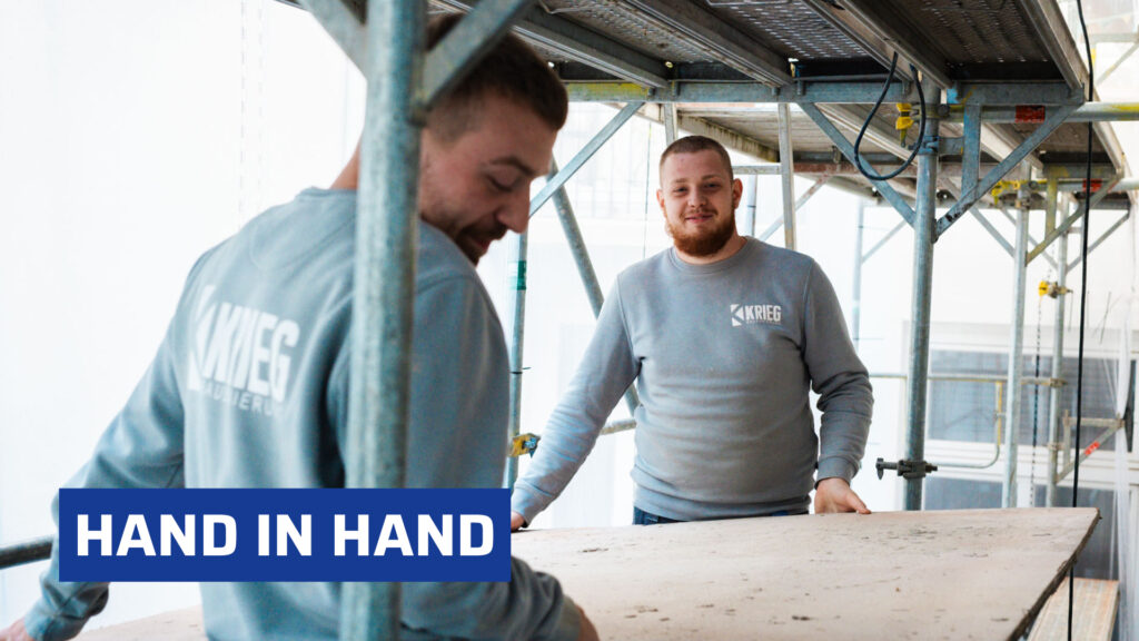 Zwei Handwerker von Krieg Bausanierung arbeiten gemeinsam auf einem Gerüst und tragen graue Pullover mit Firmenlogo; im Vordergrund ist der Schriftzug "HAND IN HAND" eingeblendet.
