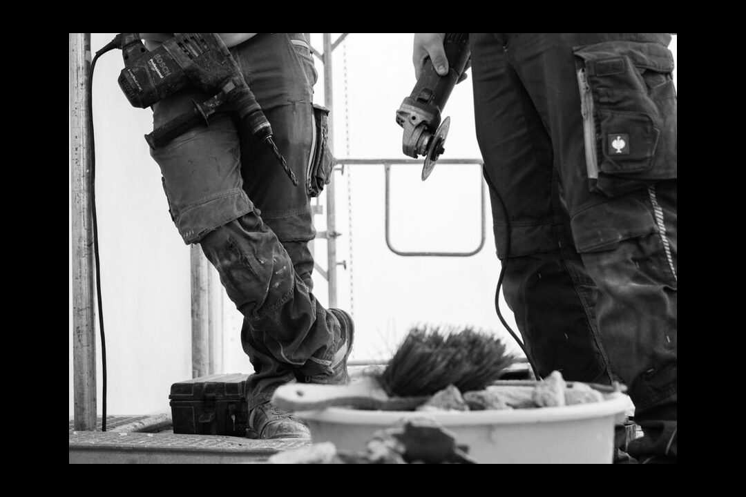 Schwarz-Weiß-Detailaufnahme zweier Handwerker auf einem Gerüst; zu sehen sind die Arbeitshosen und Werkzeuge wie ein Bohrhammer und ein Winkelschleifer.