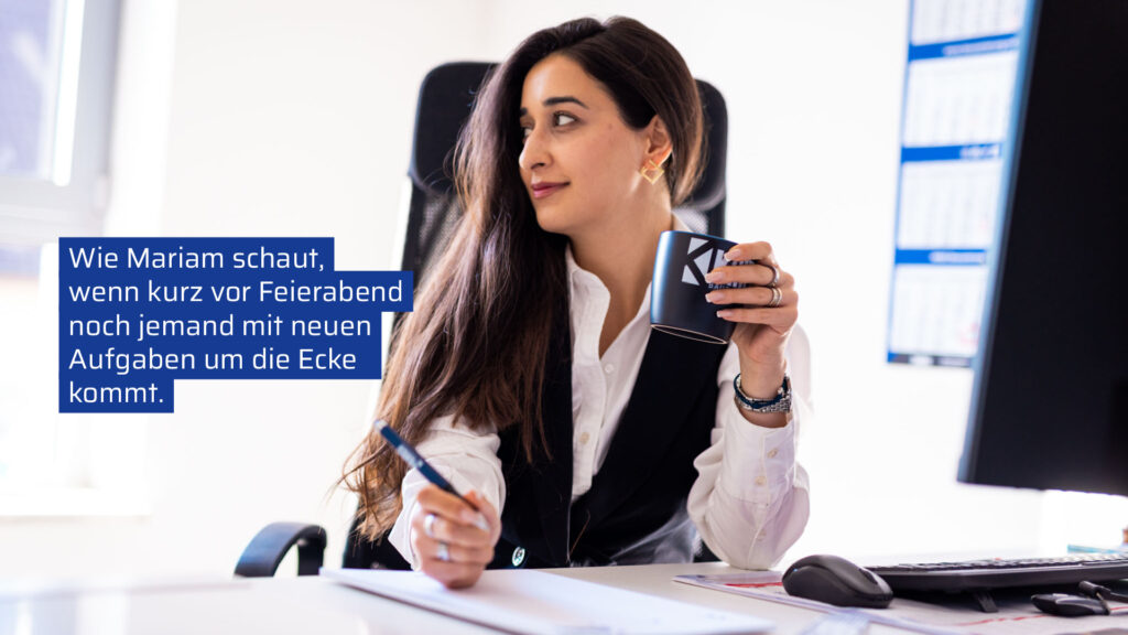 Eine Frau namens Mariam sitzt am Schreibtisch in einem Büro und blickt mit einem leicht skeptischen, aber schmunzelnden Ausdruck zur Seite. Sie hält eine Kaffeetasse in der einen Hand und einen Stift in der anderen. Links im Bild befindet sich ein blauer Textblock mit der Aufschrift: „Wie Mariam schaut, wenn kurz vor Feierabend noch jemand mit neuen Aufgaben um die Ecke kommt.“