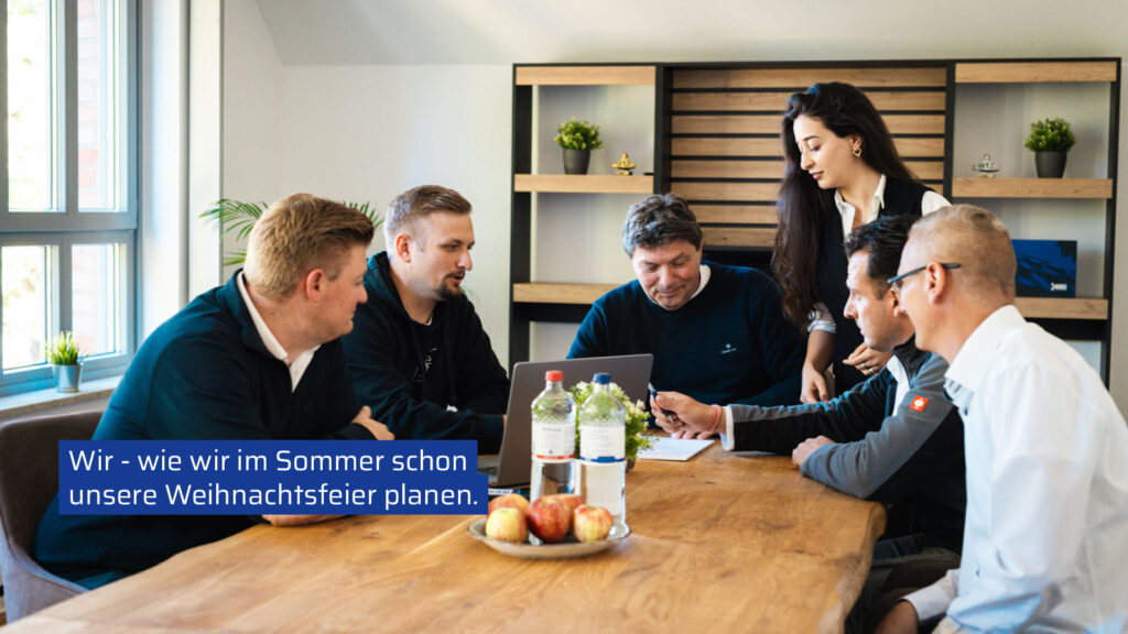 Sechs Teammitglieder der Krieg Bausanierung sitzen und stehen gut gelaunt um einen großen Holztisch im Büro. Auf dem Tisch stehen Äpfel, Wasserflaschen und ein Laptop. Ein eingeblendeter blauer Text lautet: „Wir - wie wir im Sommer schon unsere Weihnachtsfeier planen.“ Die Gruppe wirkt konzentriert und bespricht die frühzeitige Projektplanung für das Winterfest.