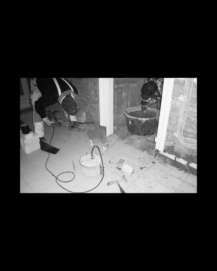Schwarz-Weiß-Foto von Bauarbeiten im Innenbereich: Zwei Bauarbeiter führen Stemmarbeiten mit Werkzeug aus und mischen Mörtel in einem Kübel an.   