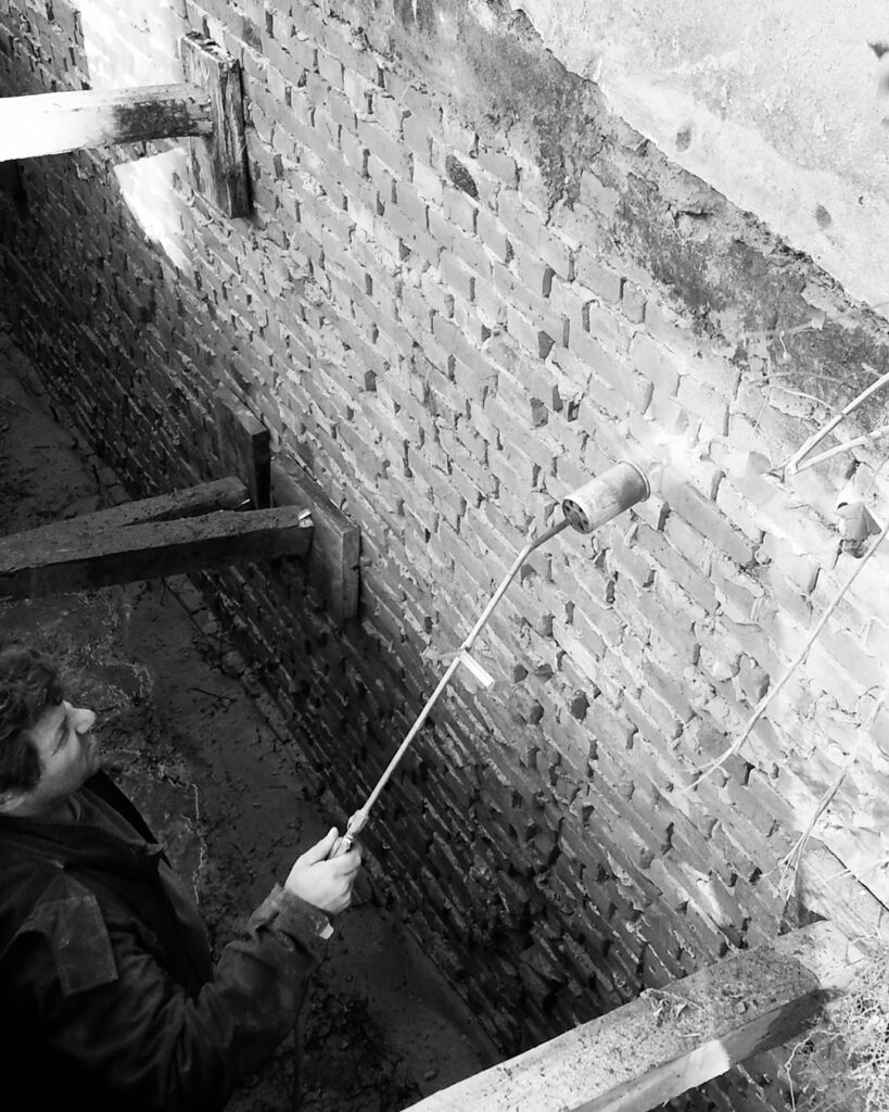 Schwarz-Weiß-Nahaufnahme eines Handwerkers, der mit einem Gasbrenner Arbeiten zur Trockenlegung und Abdichtung an einer freigelegten Backsteinwand durchführt.   