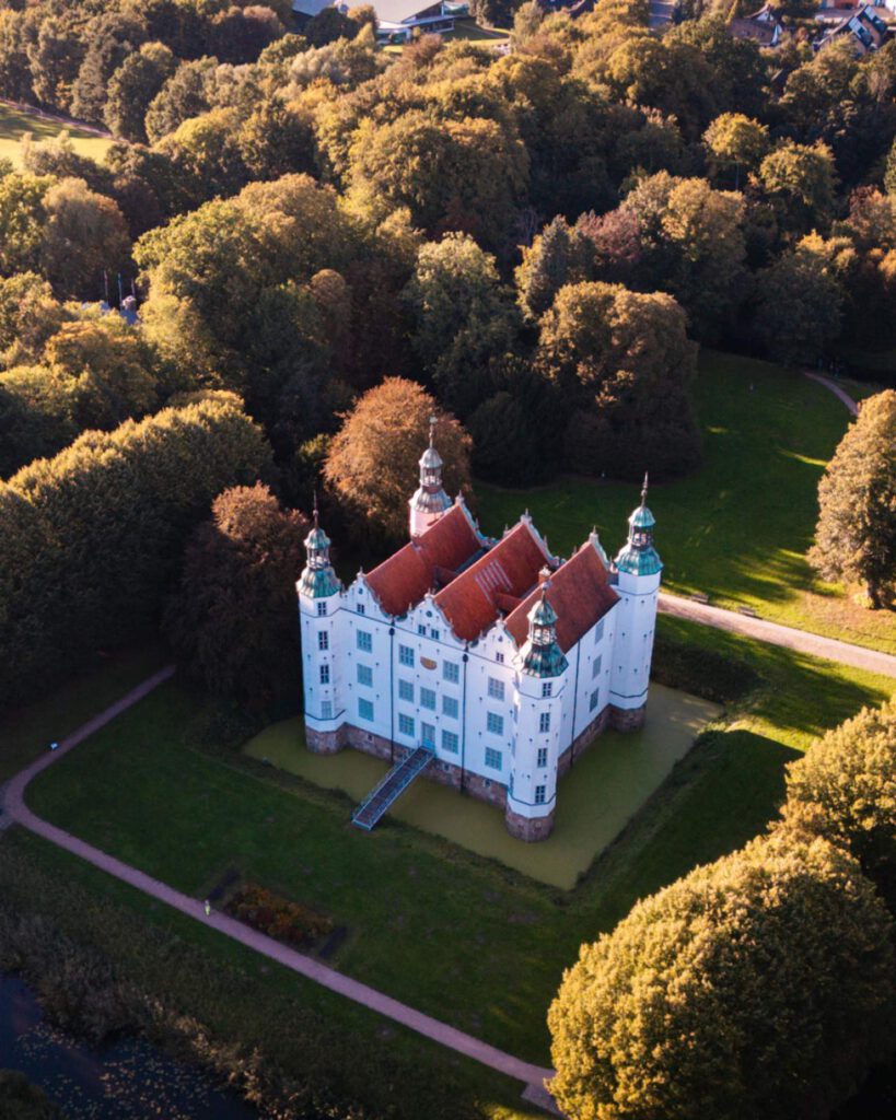 Luftaufnahme von Schloss Ahrensburg, umgeben von einem Wassergraben und herbstlichen Bäumen im Sonnenlicht.