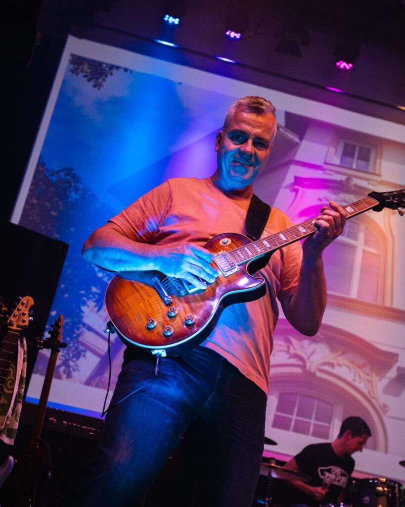 Ein lächelnder Gitarrist spielt auf einer Sunburst-E-Gitarre auf der Bühne vor einer hell beleuchteten Leinwand.   