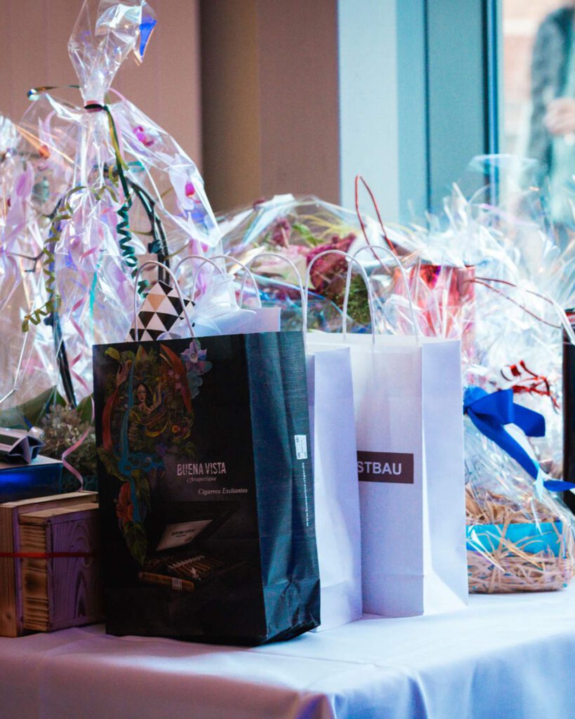 Verschiedene Geschenke und Präsente auf einem weiß gedeckten Tisch, darunter Papiertragetaschen und folierte Präsentschalen.
